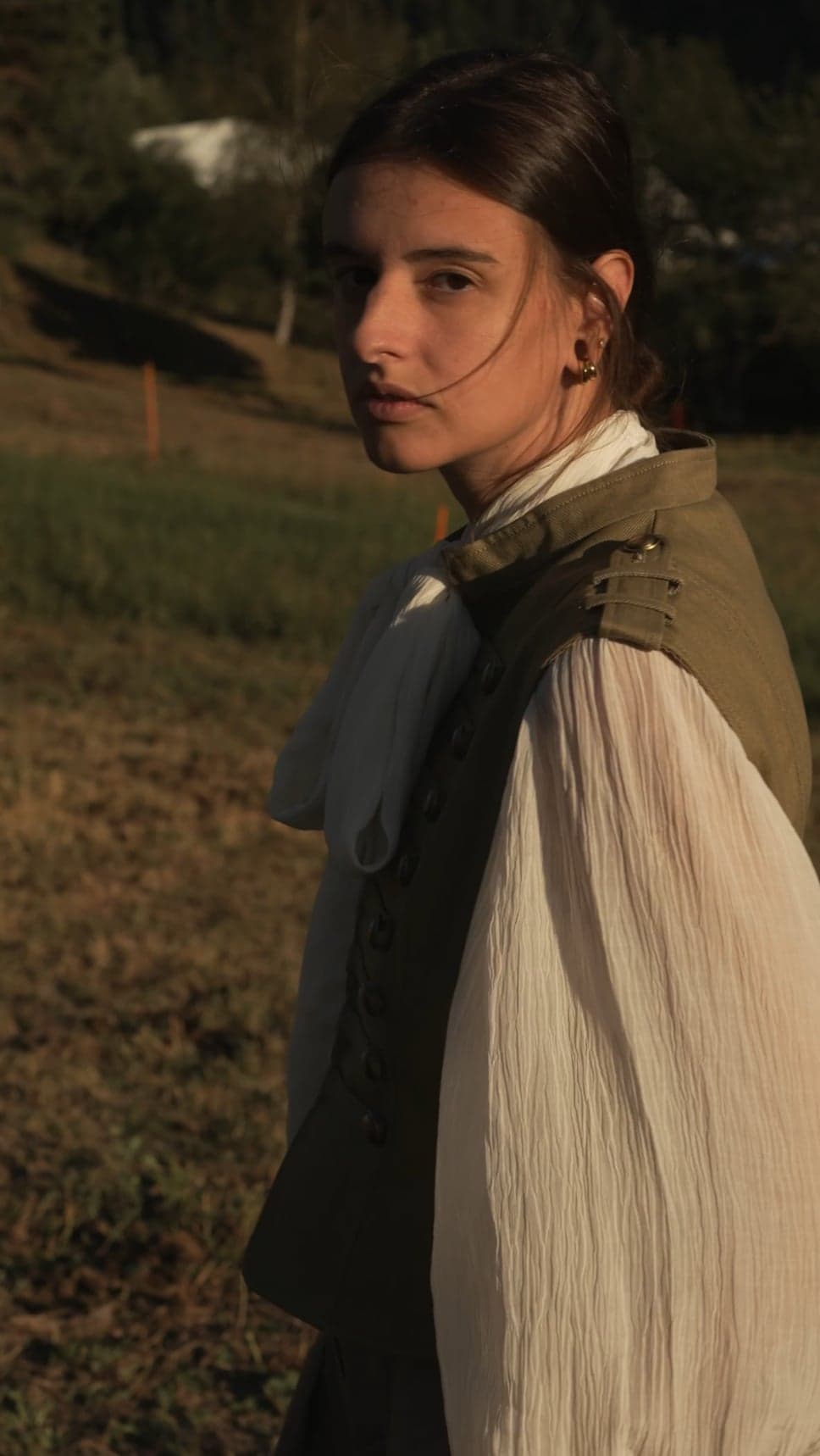 Woman in a white blouse and olive waistcoat looking back over her shoulder in evening light